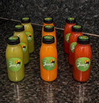 Six bottles of juice from 'The Juice Box' arranged on a dark granite countertop. The bottles contain orange, red, and green juices and are topped with black caps, each featuring a green circular label with the brand logo.