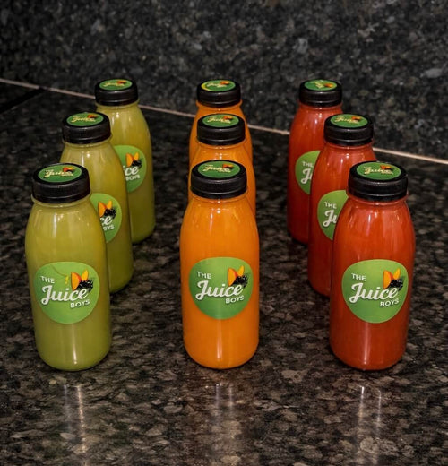 Six bottles of juice from 'The Juice Box' arranged on a dark granite countertop. The bottles contain orange, red, and green juices and are topped with black caps, each featuring a green circular label with the brand logo.