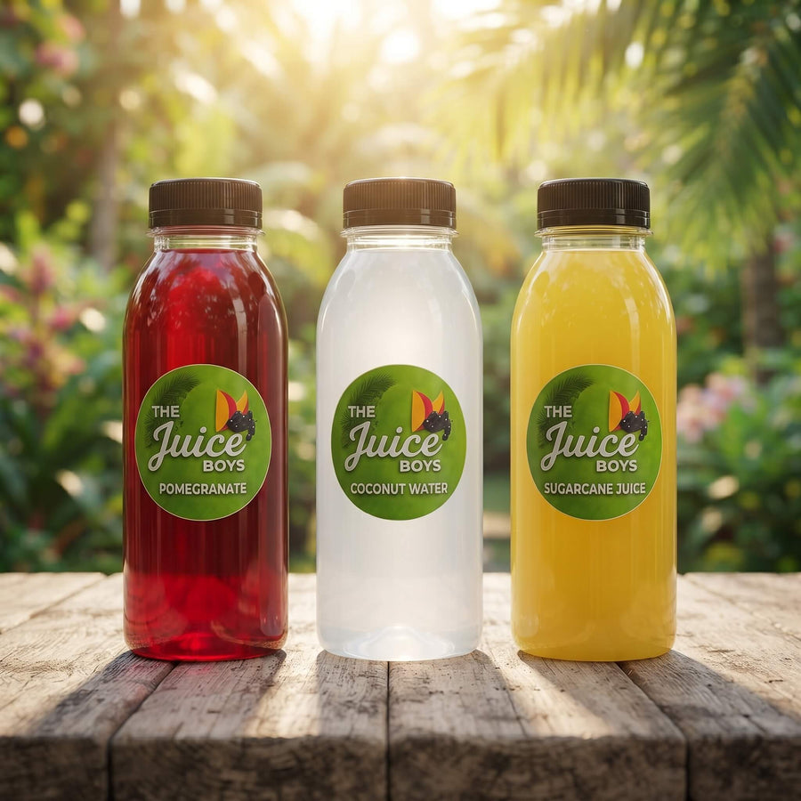 Three plastic juice bottles labeled 'THE Juice Boys' stand on a wooden table outdoors. The bottles contain red pomegranate juice, clear coconut water, and yellow sugarcane juice, with a blurred green garden background.