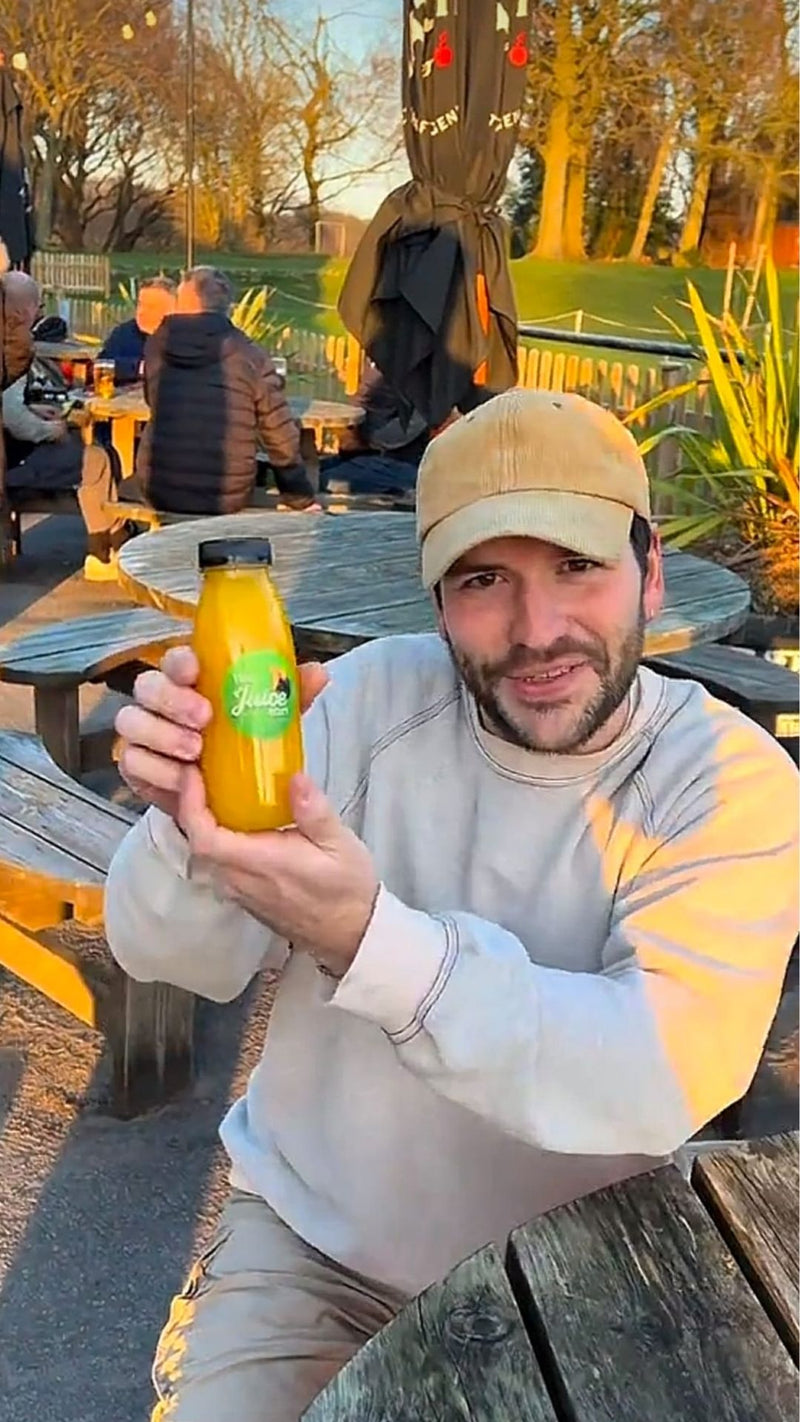 A man wearing a beige cap and a light gray sweatshirt holds up a bottle of orange juice with a green label at an outdoor patio table during golden hour.