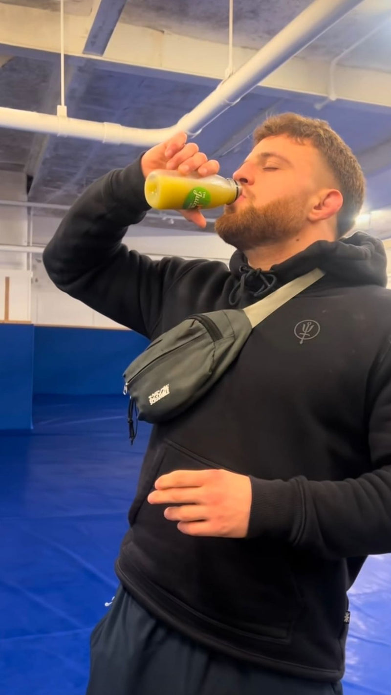 A man in a black hoodie drinking from a green bottle, wearing a black crossbody fanny pack with a white logo, in an indoor gym setting with blue mats.