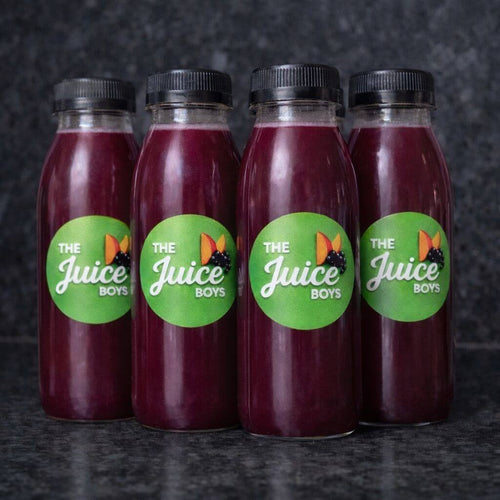 Four clear plastic juice bottles filled with deep purple-red juice, each with a black cap and a round green label reading 'The Juice Boys' with a butterfly graphic, arranged on a dark textured surface.