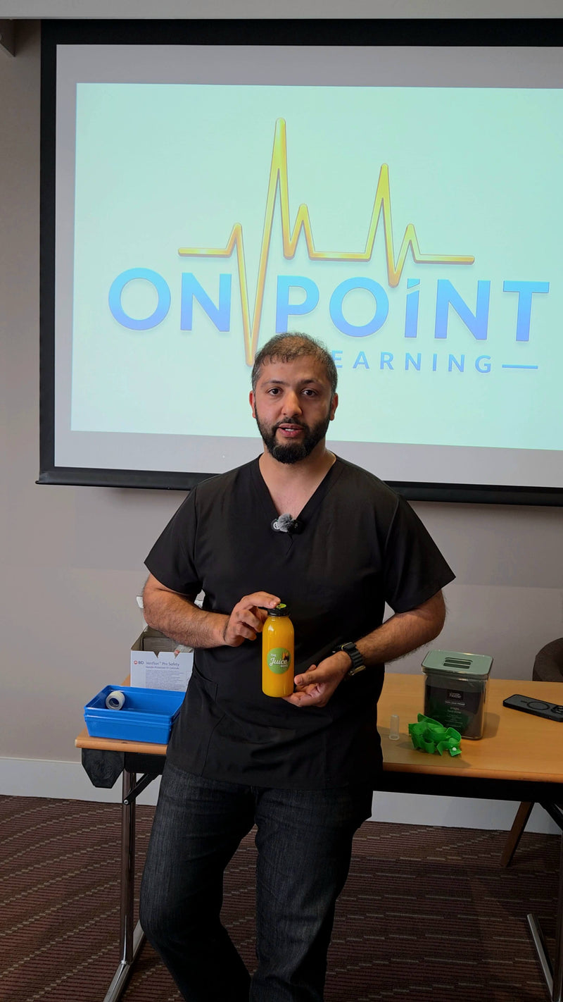 A man in black scrubs stands in front of a projection screen displaying the 'ON POINT LEARNING' logo, holding a yellow Juice Boys bottle in a presentation or training room setting.