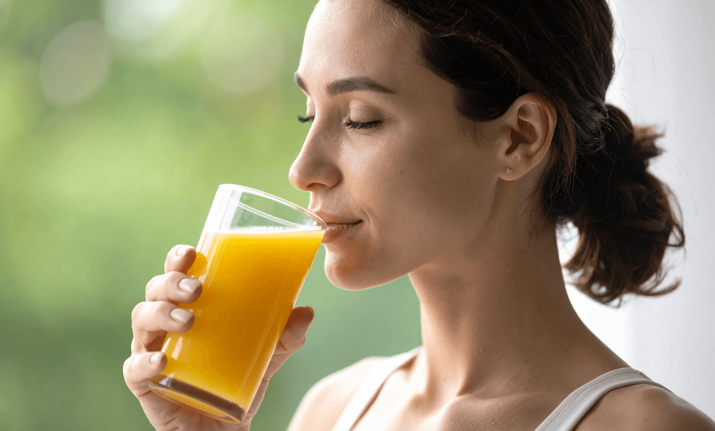 A woman with her hair in a low bun sipping orange juice from a clear glass, shown in a close-up profile shot against a soft, blurred green and white background.