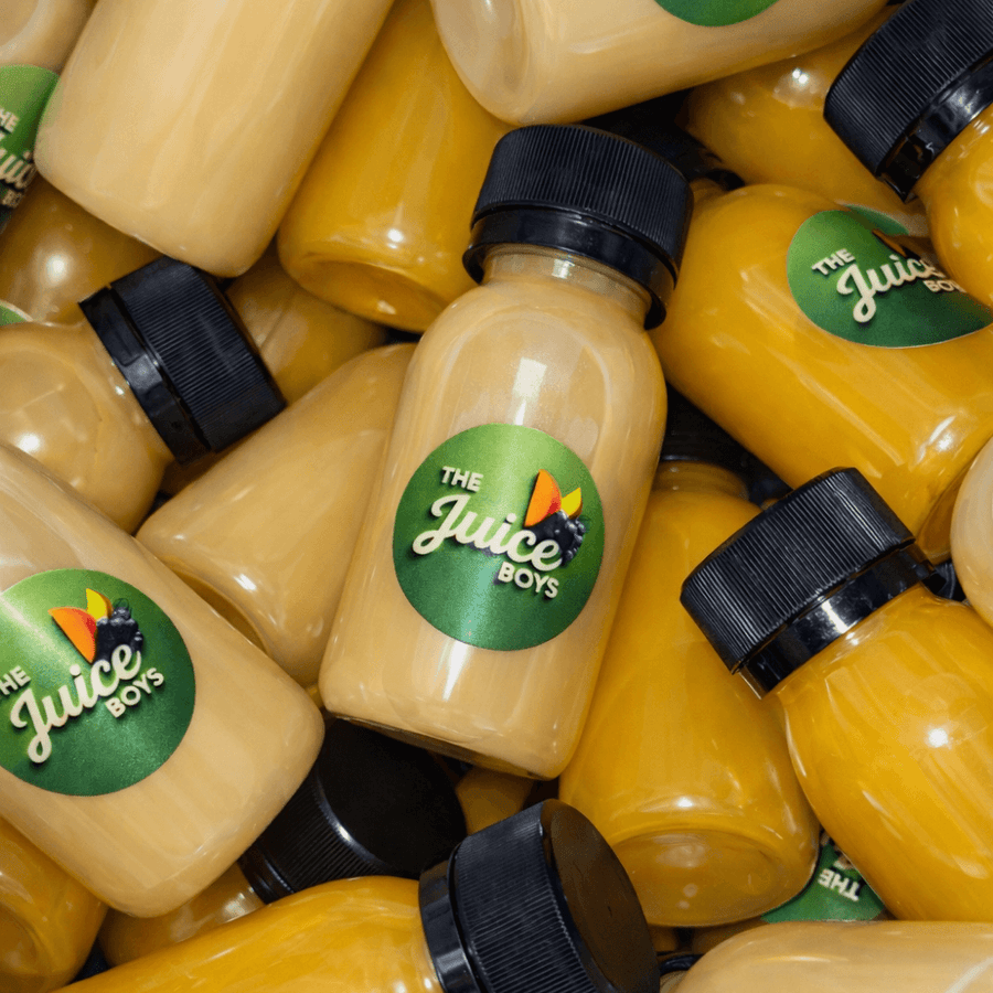 A pile of Juice Boys juice bottles, showing both the yellow-orange apple juice and a lighter yellow-beige juice, with green circular labels featuring the 'The Juice Boys' logo.